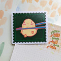 Die-cut vinyl cute sticker of a round yellow chick being held up by wooden chopsticks, cute food-themed illustration.